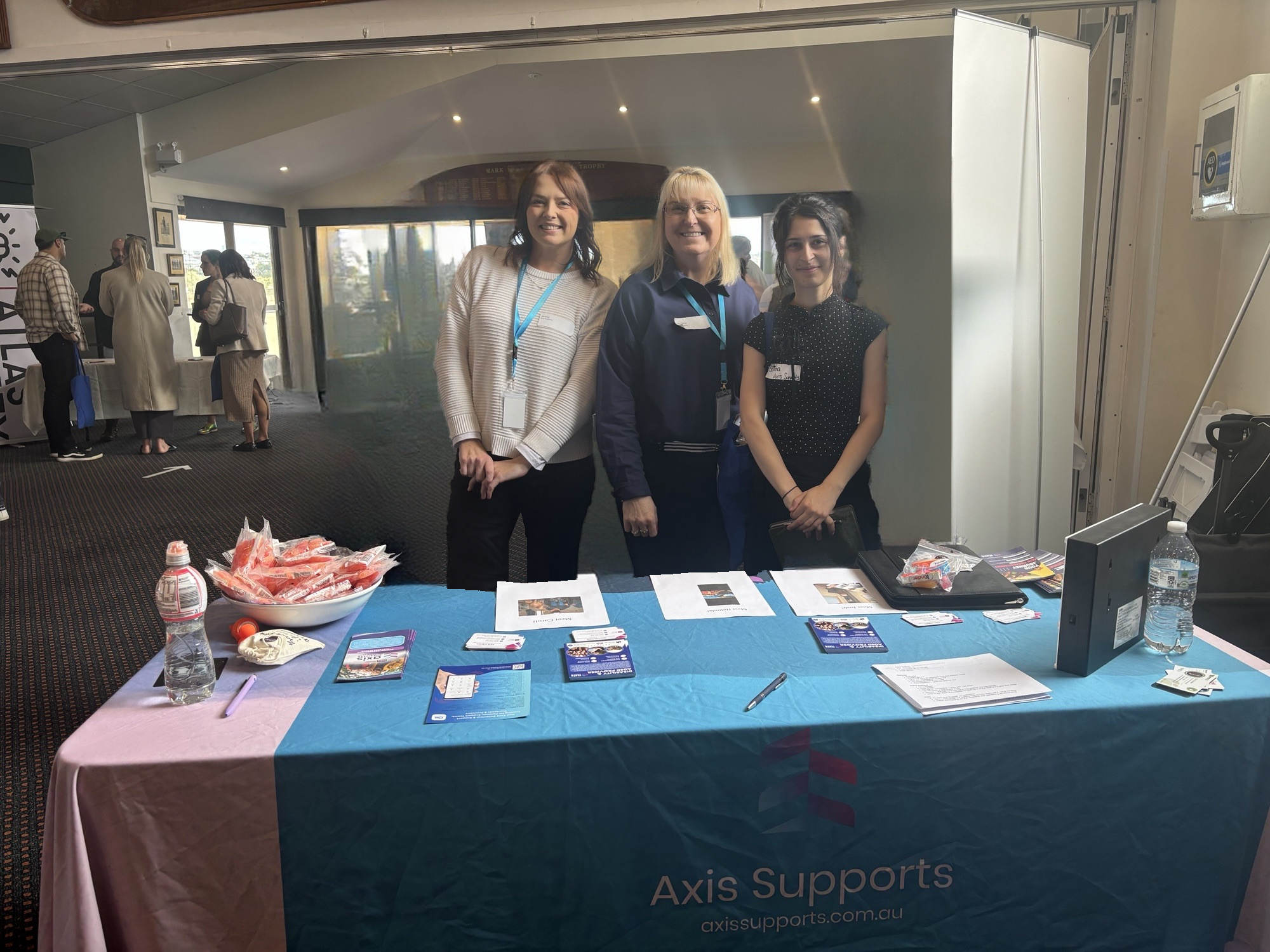 Three AXIS Supports team members smiling behind a community event stall